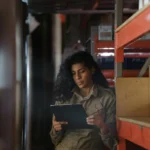 Petugas checker gudang wanita memegang clipboard memeriksa inventaris di antara rak penyimpanan